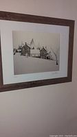 Framed print showing winter scene with stone buildings and snow, signed by John Shea