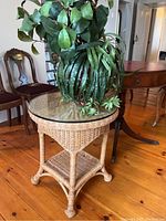 Wicker round side table with clear glass top and faux plant in decorative vase on top.