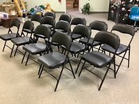 Front-side angled view of the 14 arranged folding chairs showing their black padded seats and backs and metal frames.