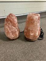 Two pink Himalayan salt lamps on wooden bases, placed on a carpeted floor, showing size and electrical cords.