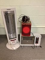 Overall lot view showing three different vintage indoor heaters: a tall white tower heater, a boxed GE Heat Lamp Kit with cage, and a small white metal heater.