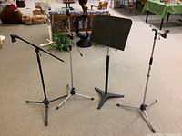 Overview of three microphone stands (two silver, one black) and one black music stand, all metal and height adjustable
