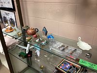 Wide view of assortment on glass shelf, including teapots, glass dolphin, small trinket boxes and compartment organizer with mini collectibles