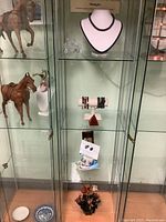 Full view of glass shelving unit displaying assorted jewelry and decorative figurines including black beaded necklace on bust and bracelets on stands.