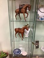 Glass cabinet shelf showing two decorative horse figurines, clear jewelry display stands, a white bust jewelry display, and small decorative plates below.