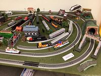 View of the train layout showing multiple model trains on oval and branching track with buildings and accessories on the green display base.
