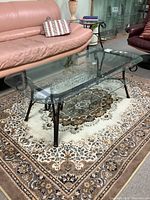 Glass coffee table showing the side view with metal frame and lower glass shelf on a patterned carpet between two pink leather sofas.