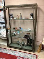 Front view of large glass display shelf/bookcase with metal frame, showing decorative edge details and multiple glass shelves with various small decorative items present (not included in sale).