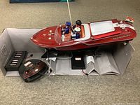 Full view of the wooden speedboat mounted on a black stand next to the black remote control and battery box inside a grey cardboard holder.