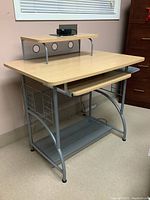 Side angle view of compact office desk showing wood laminate top, metal frame, pull-out keyboard tray, mesh lower shelf, and side magazine holders. Black device on raised shelf.