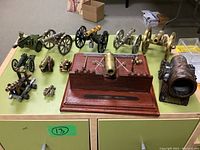 Overview of lot showing large brass cannon on wooden base and ten smaller miniature cannons arranged around it on a green tabletop
