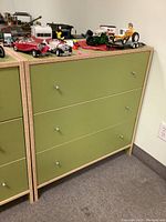 Green IKEA 3 drawer dresser with various small model cars and toys on top. Three drawers with small metal knobs, light wood framing, green drawer fronts. Shows minor wear and scuff marks.