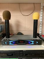 Front view of Visonic VS-1189mx dual channel wireless receiver unit with two handheld wireless microphones inserted in charging base. Microphones have colored foam windscreens (olive green and yellow). Unit is powered on with blue LED indicator lights lit.