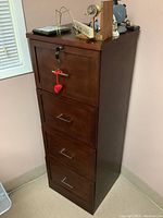 Full view of the wooden filing cabinet with four drawers, keys in lock, placed against a wall near a window, showing minor wear.