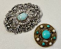 Two vintage costume brooches: a silver-tone floral scrollwork brooch with turquoise-colored stone center by Original By Robert and a round gold-tone floral and faux pearl brooch marked Germany.