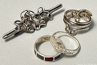 Group photo showing two rings and one brooch arranged on a light fabric background, highlighting silver finish and colored inlays on ring.