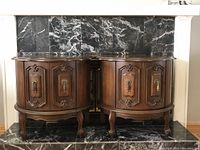 Front view of matching pair of semi-circular wooden vintage side cabinets showing ornate carved doors and brass-tone handles standing on cabriole legs on black marble hearth.