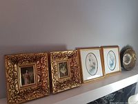 Photo showing all five framed pictures displayed on a shelf including two ornate gilded frames, two floral oval matte prints, and one round floral frame