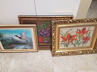 Photo showing all three framed pictures side by side on the floor: petit-point orange lilies, still life fruit, and scenic bird painting