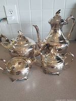 Photo showing complete set arranged on counter with white tile backsplash, including coffee pot, tea pot, sugar bowl, and creamer. Highlights the overall reflective silverplate finish and ornate handchased detailing.