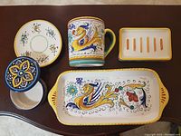 Five handpainted pottery items including mug, rectangular serving dish, small covered pot, wall plug-in holder, and small round dish displayed on table