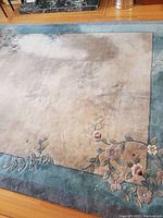 Full view of the oriental rug showing beige center and thick blue-green border with floral designs along edge and fringe intact.