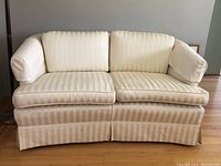Front view of a beige striped loveseat with arm protectors, showing two cushioned seats and skirted base.