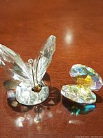 View of the Swarovski crystal butterfly and the iridescent clamshell with pearl side by side on a wooden surface, showing the clear crystal and gold face of the butterfly and colorful reflections on the clamshell.