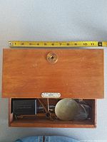Open wooden box showing brass balance scale, tray, weights and black adjustment part inside.
