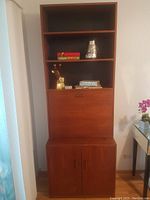 Front view of tall teak wood bookcase bar showing upper shelves with items, drop-down bar area, and bottom cabinet with doors closed