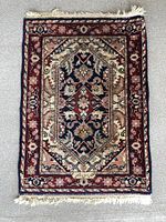 Full view of small rectangular vintage wool carpet rug on light carpeted floor showing entire rug including fringed ends.