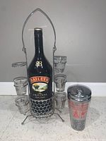 Clear view of the Baileys bottle in the stainless steel holder with five shot glasses and space for a sixth. Chrome cocktail shaker placed next to it showing printed drink recipes.