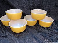 Photo showing set of six small mustard yellow bowls with white insides, arranged in two groups on black fabric.