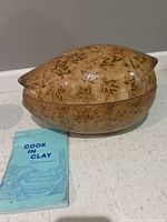 Front side view of the glazed Schlemmertopf earthenware pot placed next to a Cook in Clay recipe book, showing leaf pattern and glossy finish.