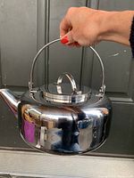 Front angled view of polished stainless steel teapot held by its handle showing rounded body and lid with loop handle.