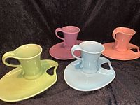 Four pastel-colored artist palette style luncheon plates each paired with a matching ceramic mug, displayed on black fabric.