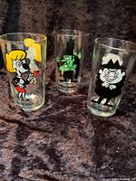 Three 1970s Pepsi collector glasses featuring Sweet Polly, Snidely Whiplash, and Boris Badenov characters, displayed on dark fabric background.