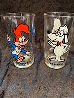 Two clear Pepsi collector glasses featuring Woody Woodpecker and Mr. Peabody cartoon characters, standing side by side on a dark fabric.
