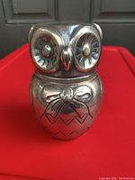 Front angled photo showing detailed silver plated owl bank with textured eyes and bow on chest, placed against a dark background.