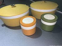 Four vintage Thermos food storage items on a surface: two large yellow food storage containers with lids and two small insulated jars in orange and olive green with white bands.