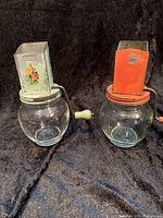 Pair of vintage nut grinders with screw-in glass jar bases shown from front angles on black fabric background.