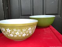 Two Pyrex mixing bowls on red surface in front of door, showing the Crazy Daisy patterned bowl in front and the plain olive green bowl behind.