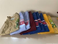 Photo of 7 folded tops and t-shirts in various colors showing graphic prints stacked horizontally on a flat surface.