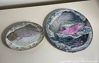 Both porcelain fish plates shown side by side on a flat surface, showcasing their size difference and painted designs of fish with blue and pink hues.