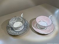 Two teacup and saucer sets in pink and grey, showing swirly leaf patterns and scalloped edges.