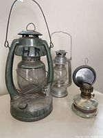 Three antique oil lanterns arranged together. Large greenish metal Embury Air Pilot lantern prominent in front left, medium silver metal lantern behind it and smaller clear glass lantern in front right.