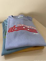 Stack of three folded large women's nice+smooth t-shirts showing the top blue-purple shirt with thumbs-up graphic and part of other two shirts underneath.