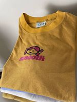 Mustard yellow T-shirt with stylized girl face and 'smooth' logo visible in close-up.