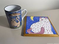 Photo of a ceramic mug with blue background decorated with cat and bird artwork, and a square ceramic trivet featuring a white spotted cat in a night scene with moon, framed in wood.
