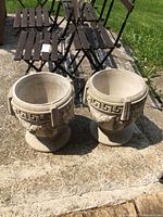 Pair of off-white concrete urns placed outdoors on concrete. Both urns have classical Greek key design near the rim and decorative grape clusters on the body.
